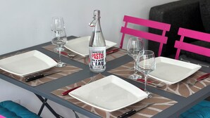 Una mesa de comedor dispuesta con platos blancos, cubiertos, vasos, una botella de agua y una botella de pastis.