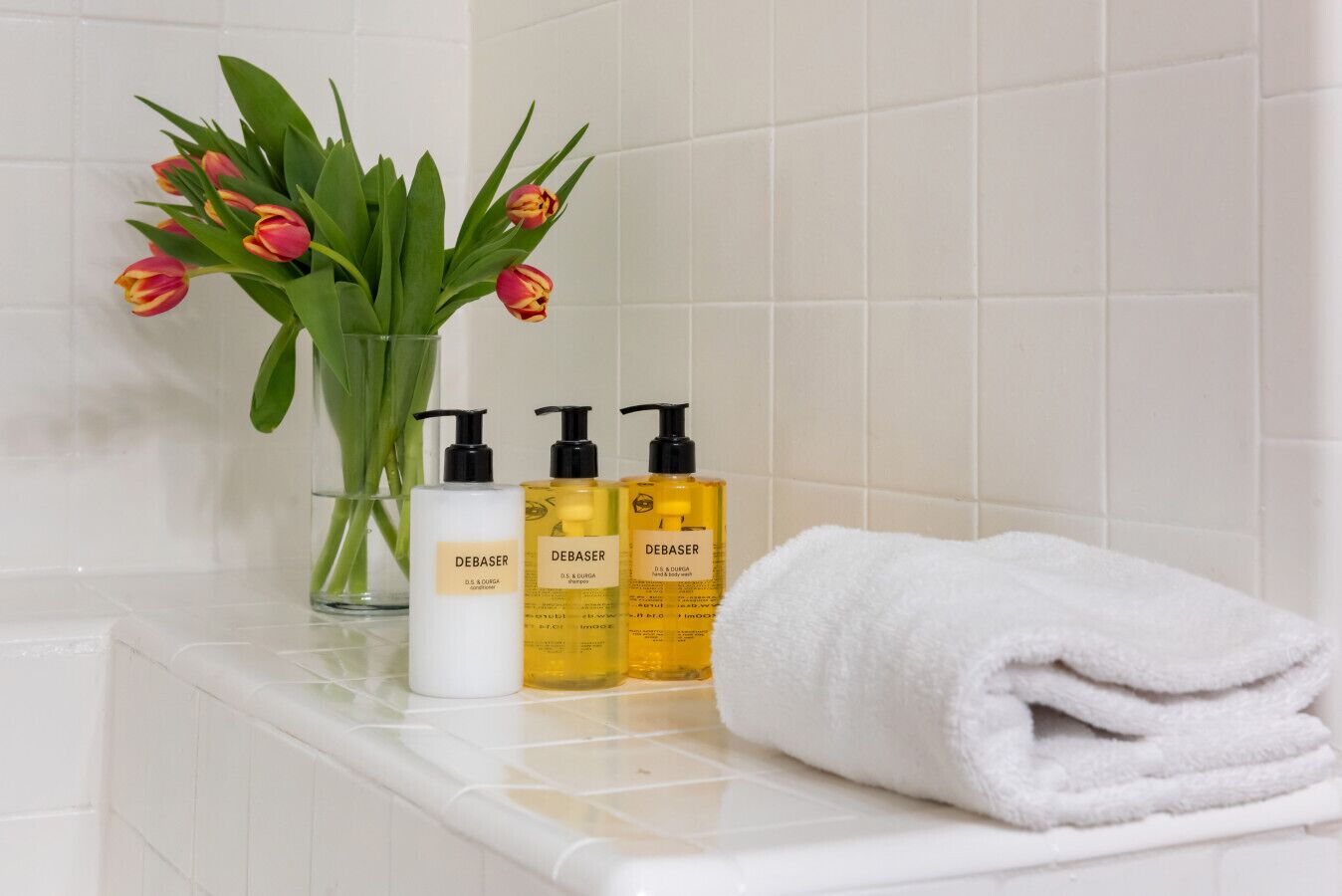 A bathroom with a white tiled wall, a white towel, and three bottles of Debaser shampoo.