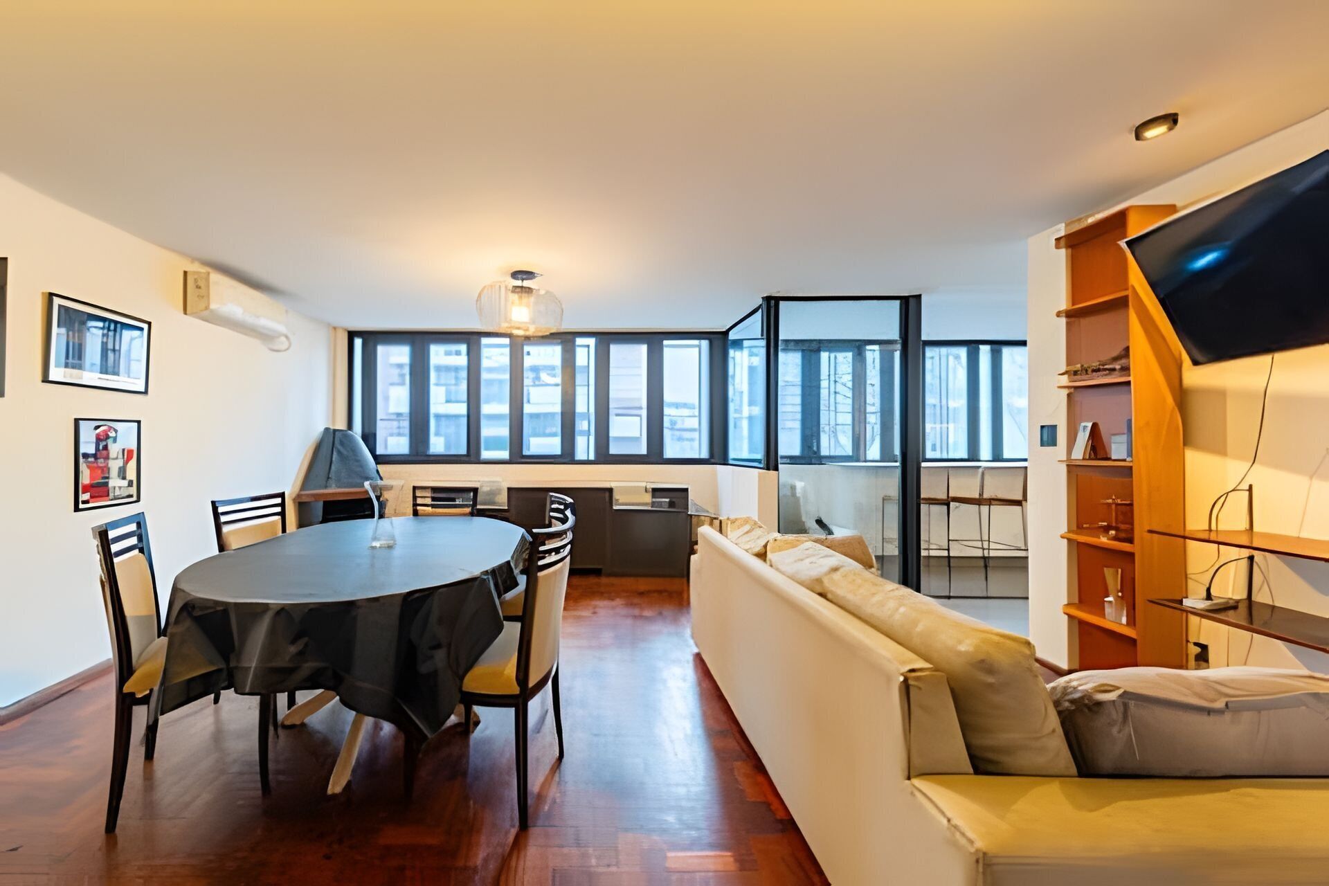 A modern living room with a dining area, large windows, a sofa, and a TV on a wooden shelf.