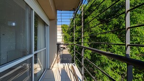 Un balcón con barandilla de metal, ventanas de vidrio y vista a árboles verdes y frondosos.
