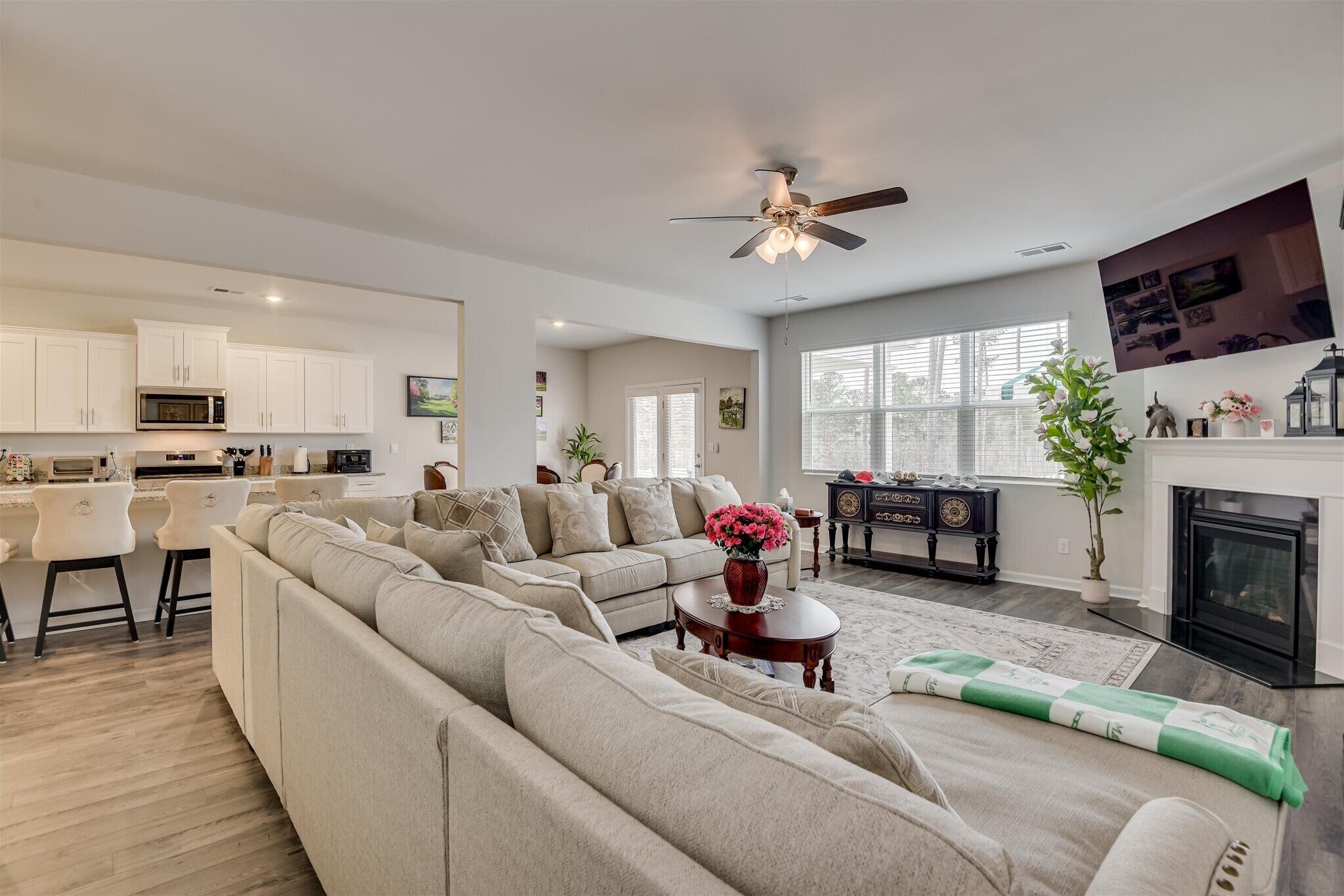 A modern living room with a large beige sectional sofa, a coffee table with a vase of flowers, a fireplace, and a kitchen area with white cabinets and a microwave.
