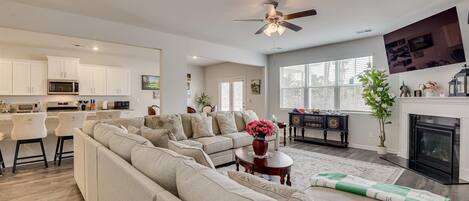 A modern living room with a large beige sectional sofa, a coffee table with a vase of flowers, a fireplace, and a kitchen area with white cabinets and a microwave.