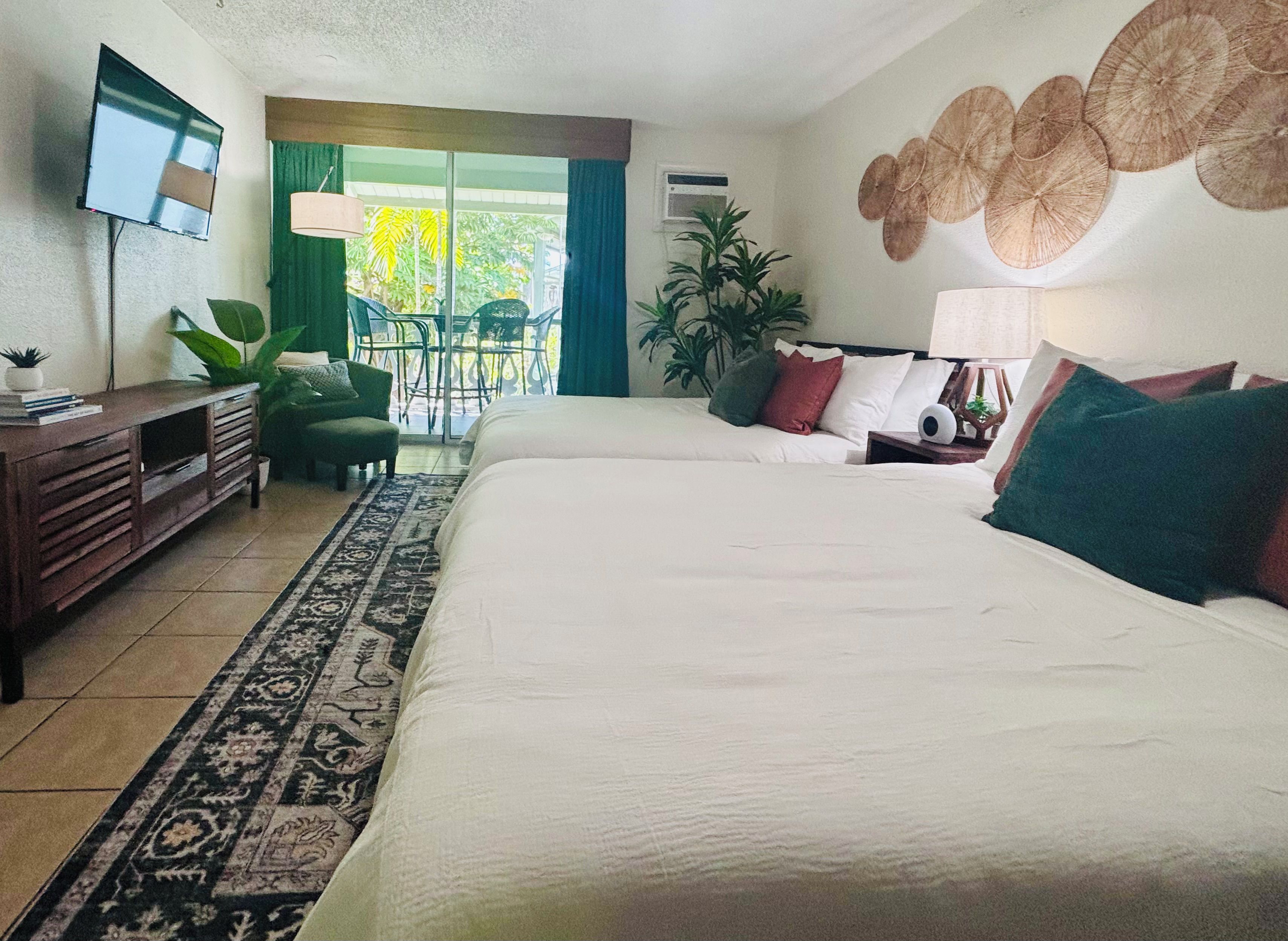 A hotel room with a bed, a desk, a chair, a TV, and a balcony with plants.