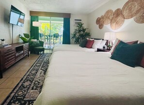 Una camera d'albergo con un letto, una scrivania, una sedia, una TV e un balcone con piante.