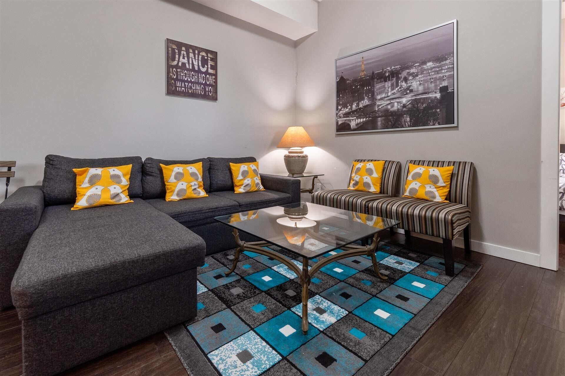 Un salon moderne avec un canapé gris, des fauteuils à rayures et une table basse en verre. Une lampe et un tableau encadré représentant un paysage urbain décorent les murs.