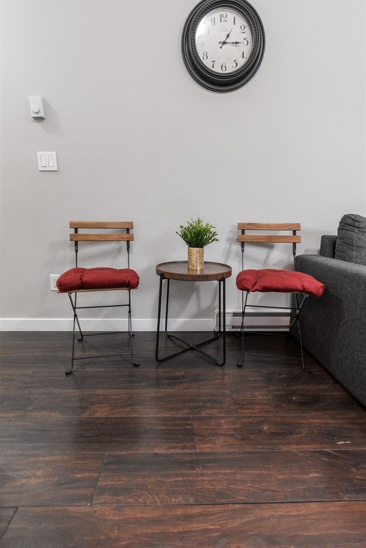A room with a dark wooden floor, two wooden chairs with red cushions, a small round table with a plant, a gray sofa, and a wall clock.