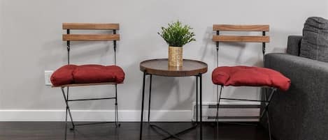 A room with a dark wooden floor, two wooden chairs with red cushions, a small round table with a plant, a gray sofa, and a wall clock.