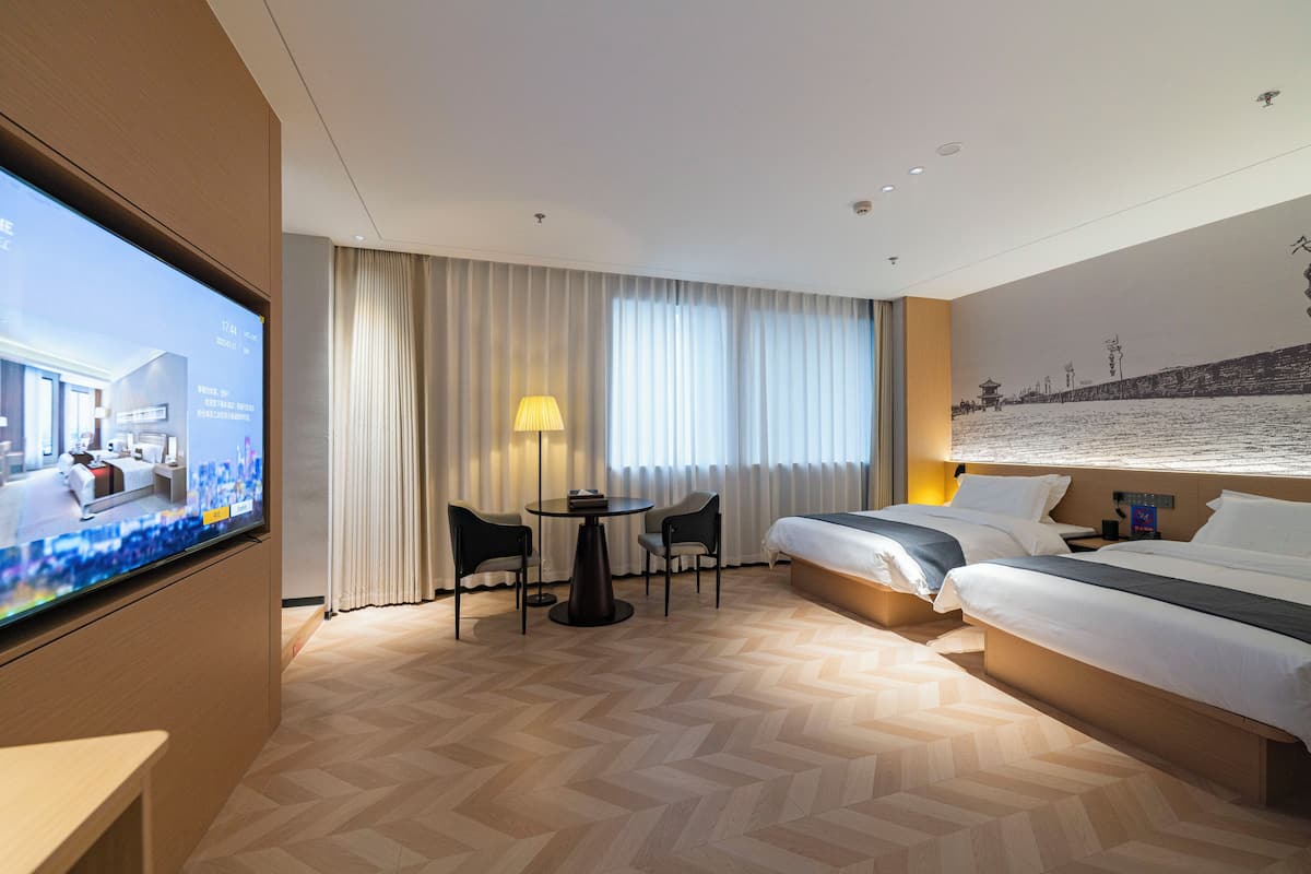 A hotel room with a bed, two chairs, a small table, a TV, and a view of a cityscape through the window.