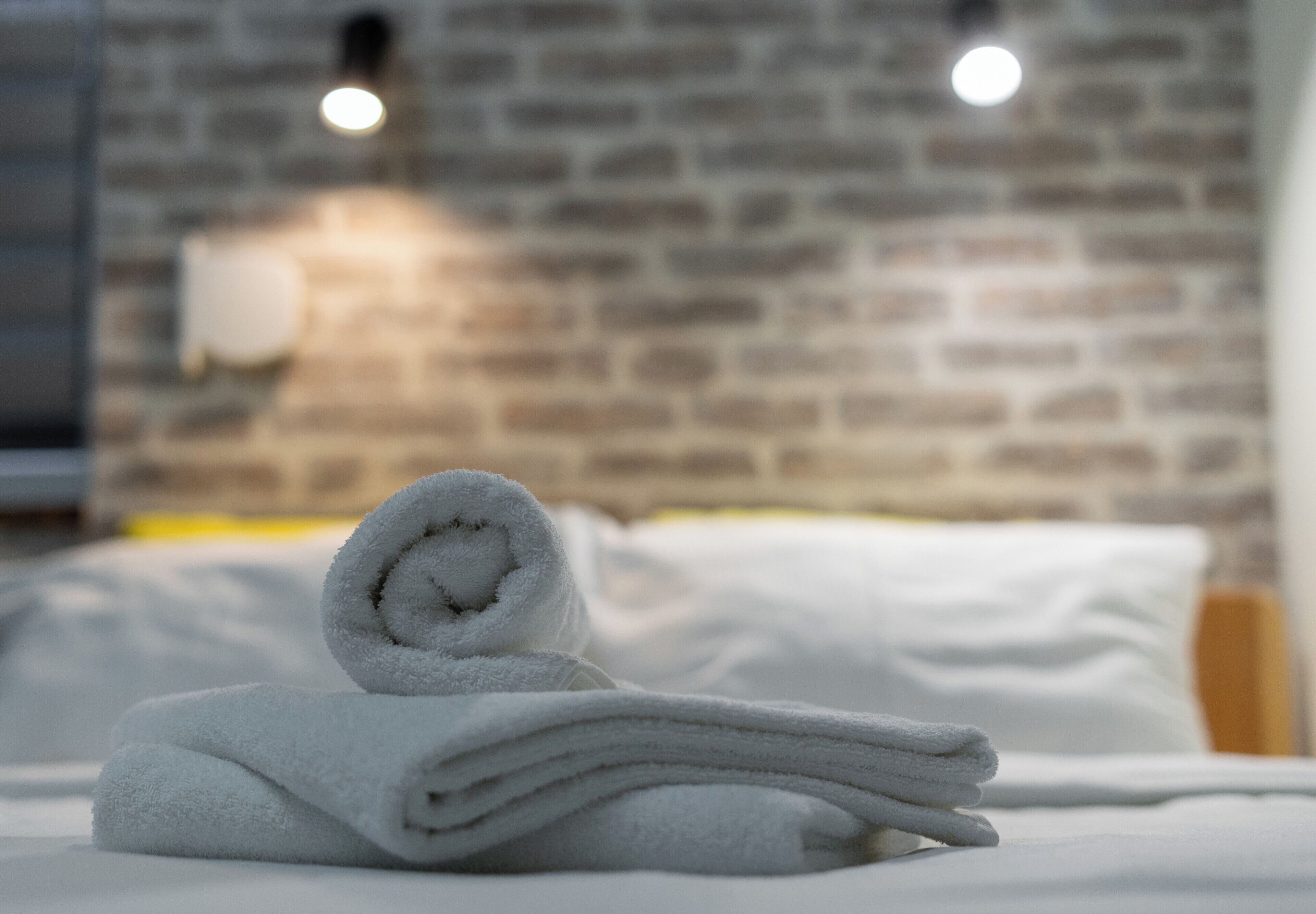 Une serviette blanche, soigneusement pliée, est posée sur un lit recouvert de draps blancs, dans une pièce dont un mur est en briques et qui est éclairée par un luminaire.