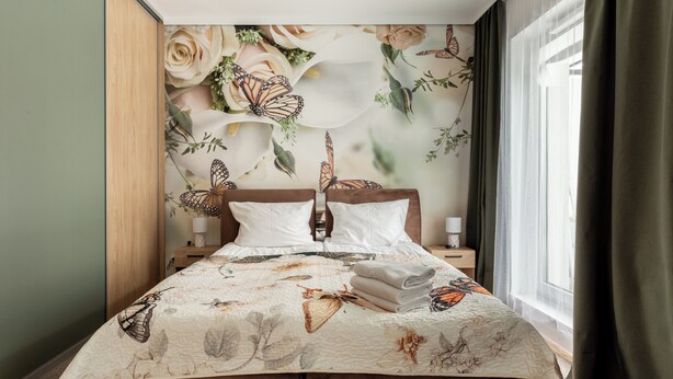 Una cama bien hecha con una colcha de motivos florales y mariposas, un cabecero de madera y una mesita de noche con una lámpara.