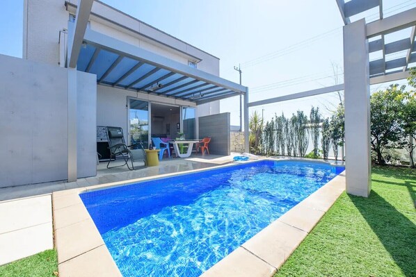 Une piscine extérieure moderne dotée d’un patio couvert, d’un espace aménagé avec des sièges et offrant une vue sur la végétation environnante.
