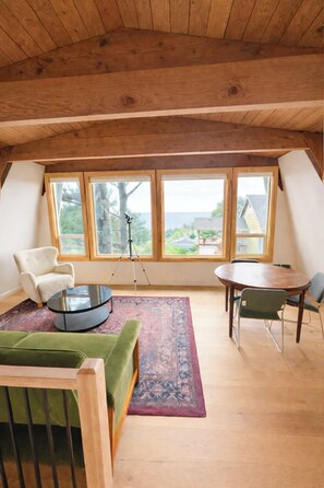 Een loft-achtige kamer met een hoge plafond, grote ramen, een ronde tafel, een groene bank en een klein rond bijzettafeltje.