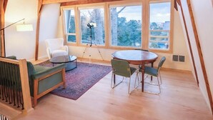 En rymlig loftvåning med högt i tak, stora fönster med utsikt över omgivningen, ett runt matbord med stolar och en mysig sittgrupp med en soffa och fåtöljer.