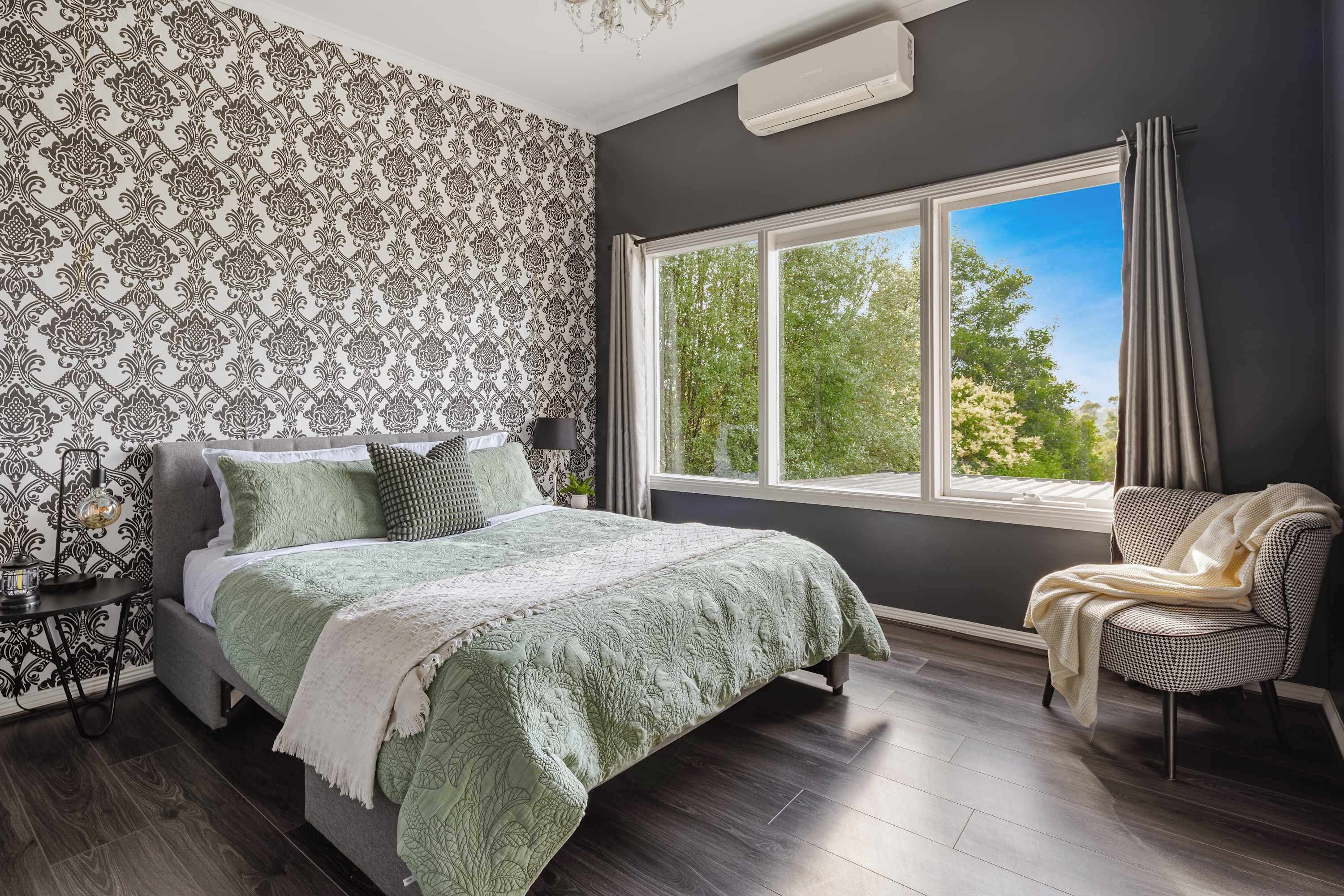 Un dormitorio con una cama grande, una silla, un ventanal con cortinas y un papel tapiz estampado.