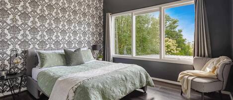 A bedroom with a large bed, a chair, a window with curtains, and a patterned wallpaper.