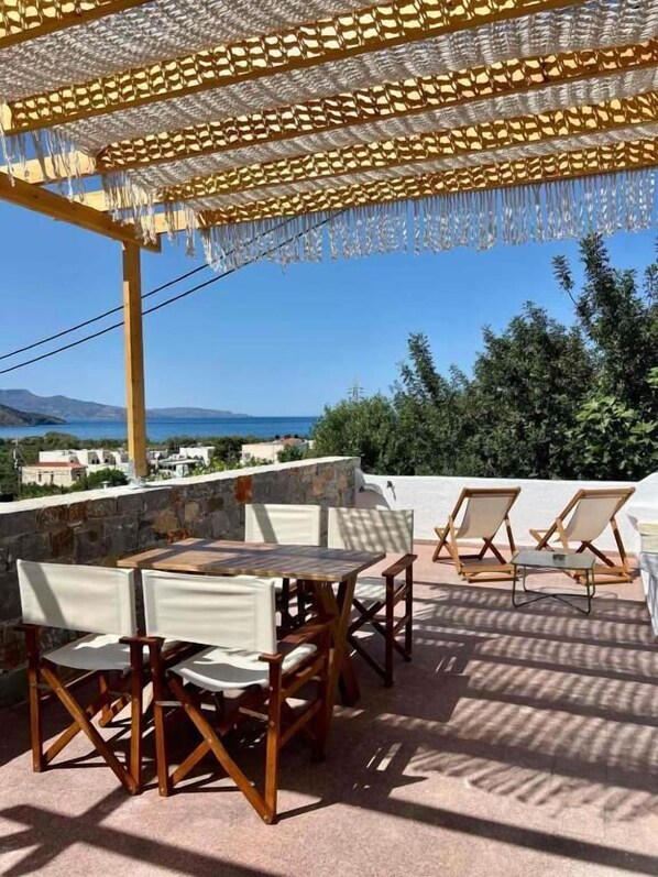 Eine Terrasse im Freien mit Tisch und Stühlen, eine Pergola mit Stoffvorhängen und Blick auf das Meer und die Hügel.