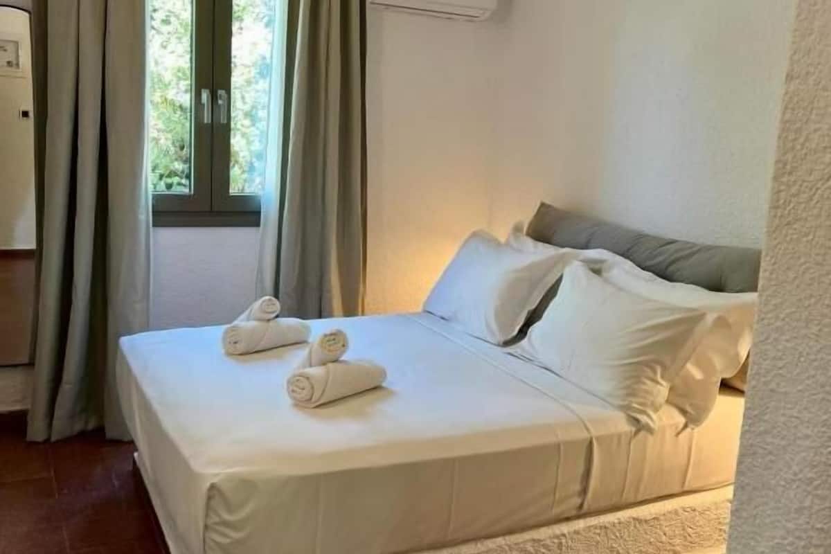 Een net opgemaakt bed met witte lakens en twee opgerolde handdoeken, een raam met gordijnen en een airconditioning op de muur.
