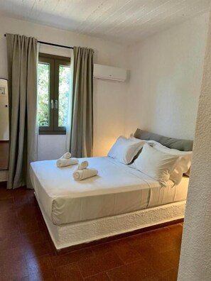 Ein ordentlich bezogenes Bett mit weißen Leinentüchern und zwei aufgerollten Handtüchern, ein Fenster mit Vorhängen und eine Klimaanlage an der Wand.