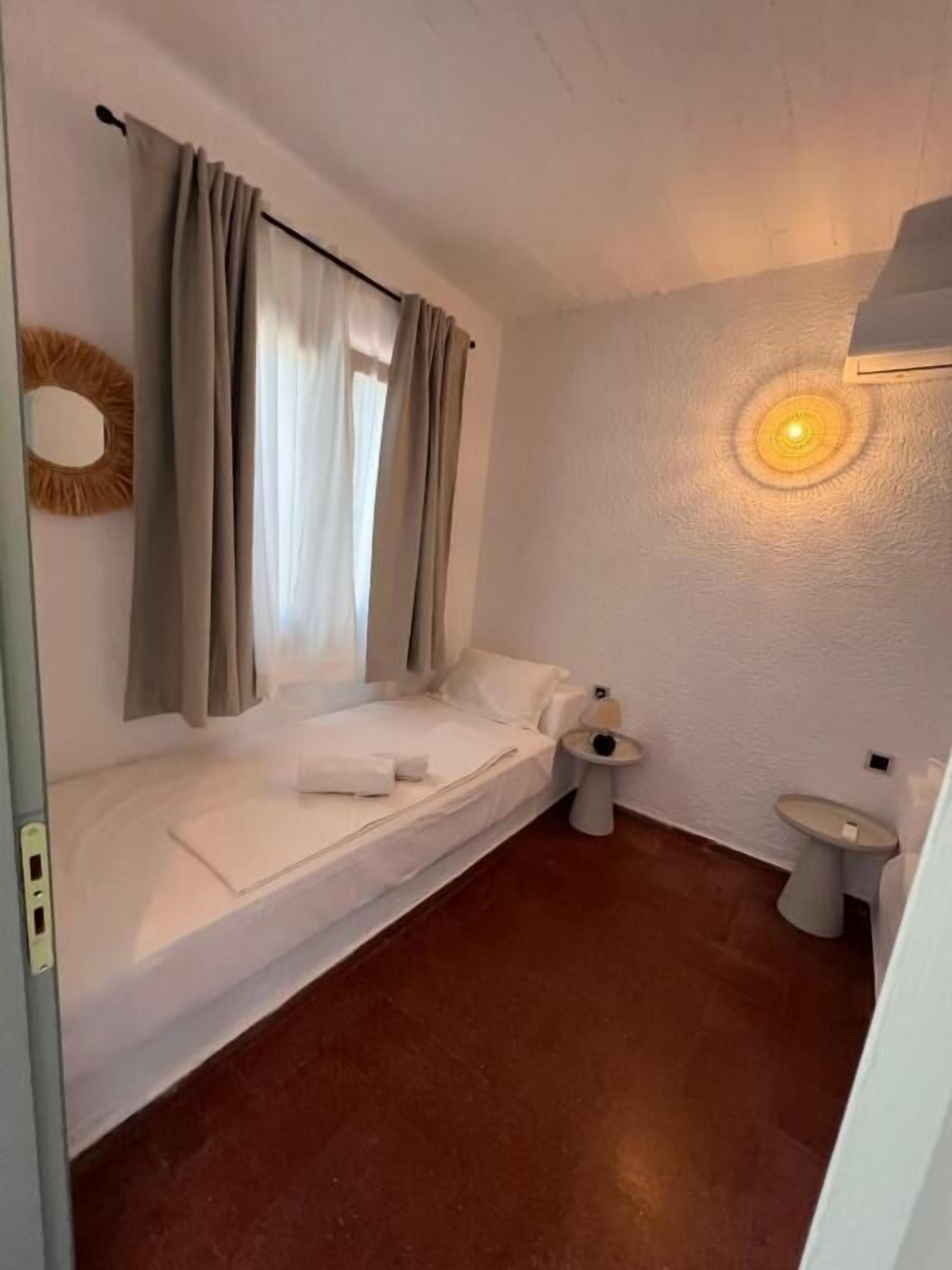 A small, single-bed room with a window, a bedside table, a wall-mounted mirror, and a toilet.