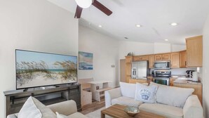 A modern living room with a flat-screen TV, a sofa, a coffee table, and a dining area with wooden cabinets and a kitchen in the background. - Walk to Beach | Pool, Deck & Easy Gulf Shores Stay (Gulf Shores)