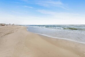 Une vaste plage de sable fin, avec des vagues douces, un ciel dégagé et des bâtiments qui se dessinent à l’horizon.