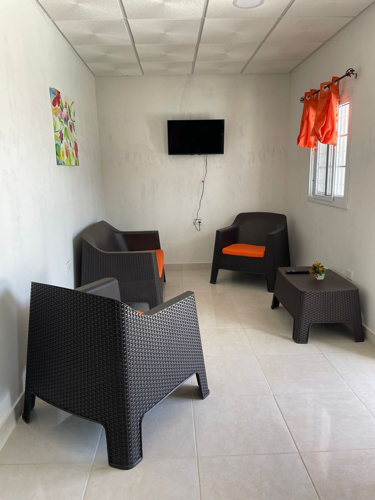Une pièce comprenant deux fauteuils en osier noir avec des coussins orange, une petite table sur laquelle est posé un vase, un téléviseur à écran plat fixé au mur et une fenêtre ornée de rideaux orange.