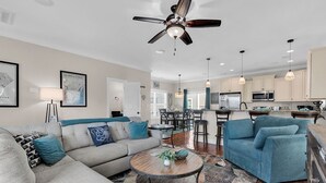 A spacious living room with a sofa, a blue armchair, and a coffee table. There is a ceiling fan, pendant lights, and a kitchen area in the background. - The Salty Mermaid - Walk to Garden City Beach (Garden City Beach)