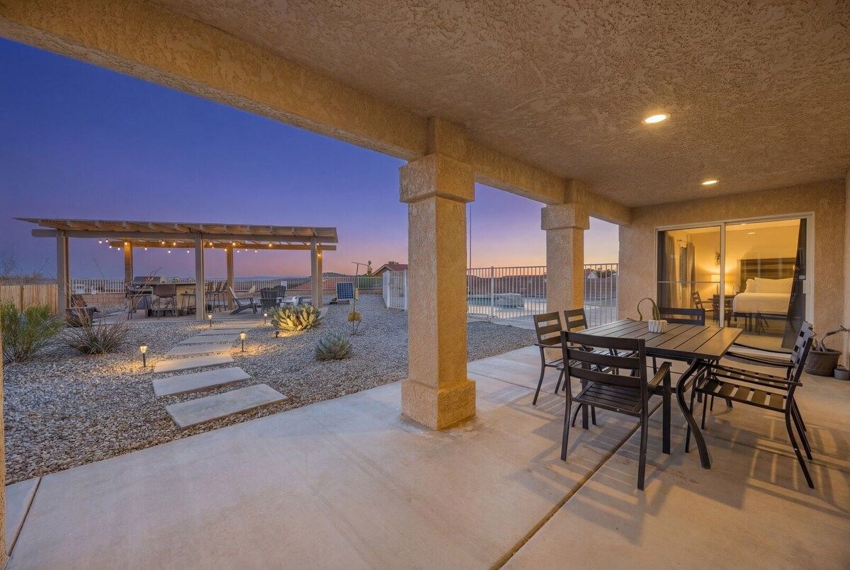 An outdoor patio area with a dining table and chairs, a pergola with string lights, and a view of the landscape.