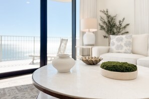 Ein runder Tisch mit einer weißen Vase und einem moosartigen Gegenstand, ein weißes Sofa mit gemusterten Kissen und eine Lampe in einem modernen Raum mit Meerblick.