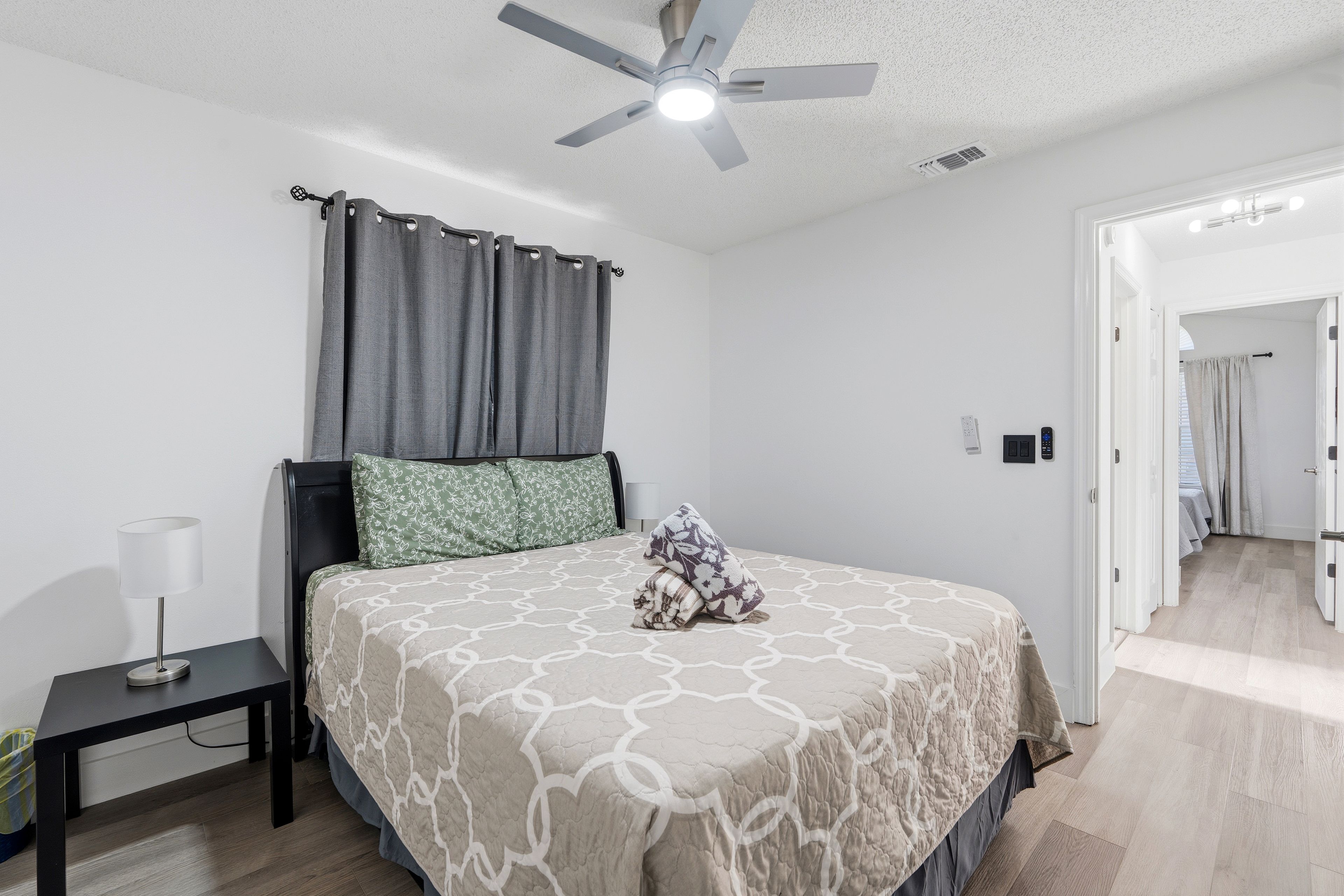A bedroom with a bed, a nightstand, a lamp, a ceiling fan, and a door leading to another room.