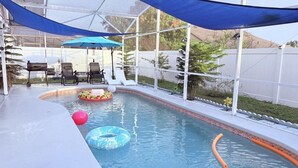 En swimmingpool med solstolar och parasoller, omgiven av ett staket och grönska.