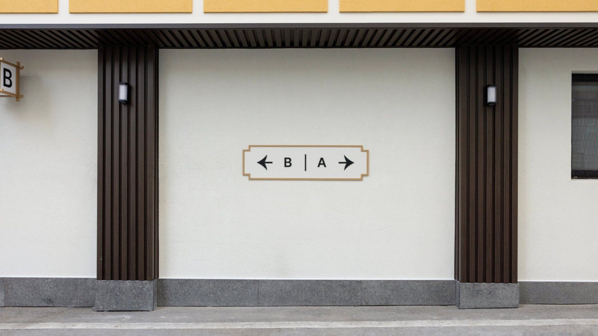 A building facade with a minimalist design, featuring a large wall-mounted sign with directional arrows and the letters "B" and "A".