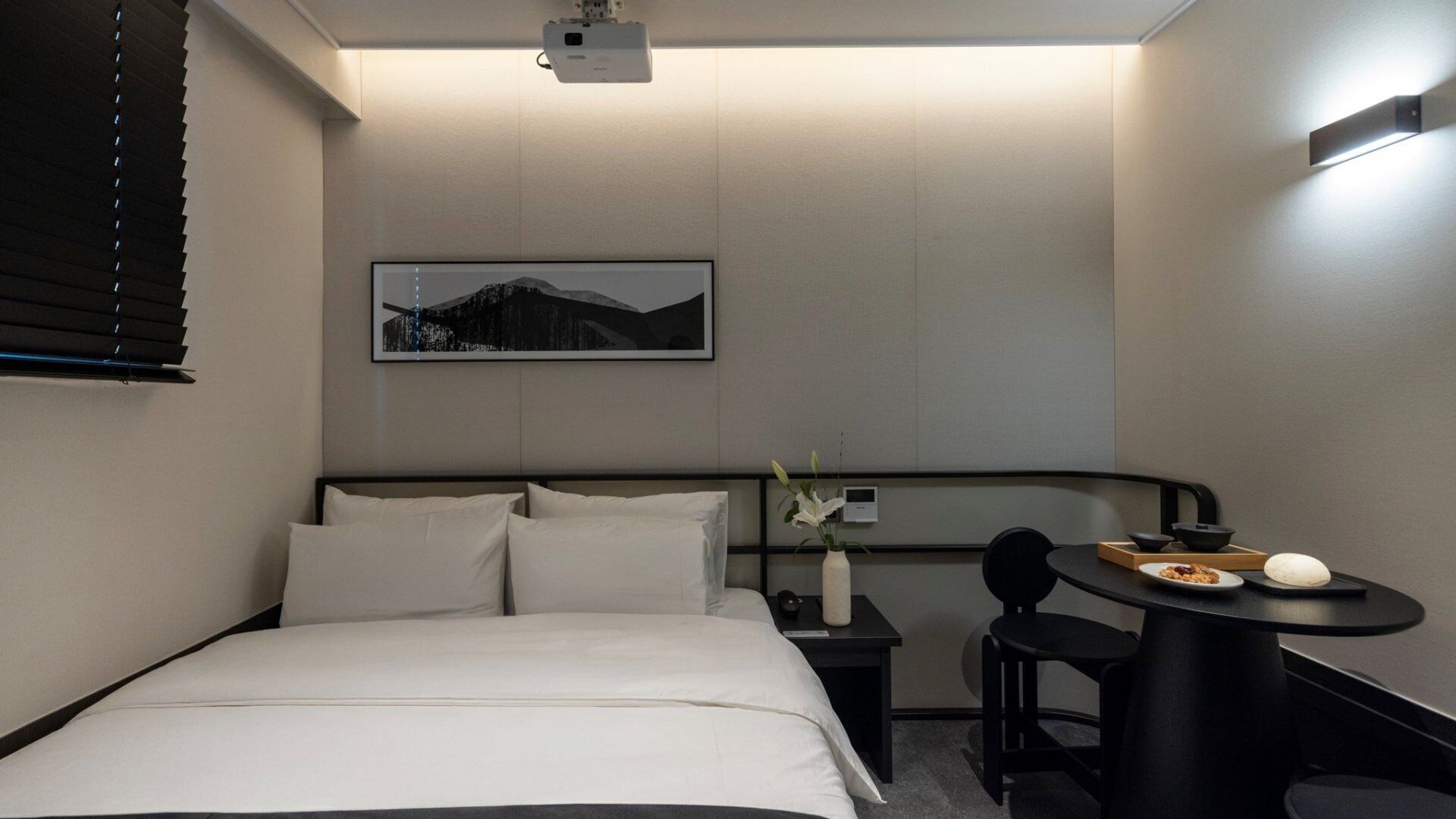 A hotel room with a bed, a small table with a bowl and a plate, a chair, a vase with flowers, and a projector on the ceiling.
