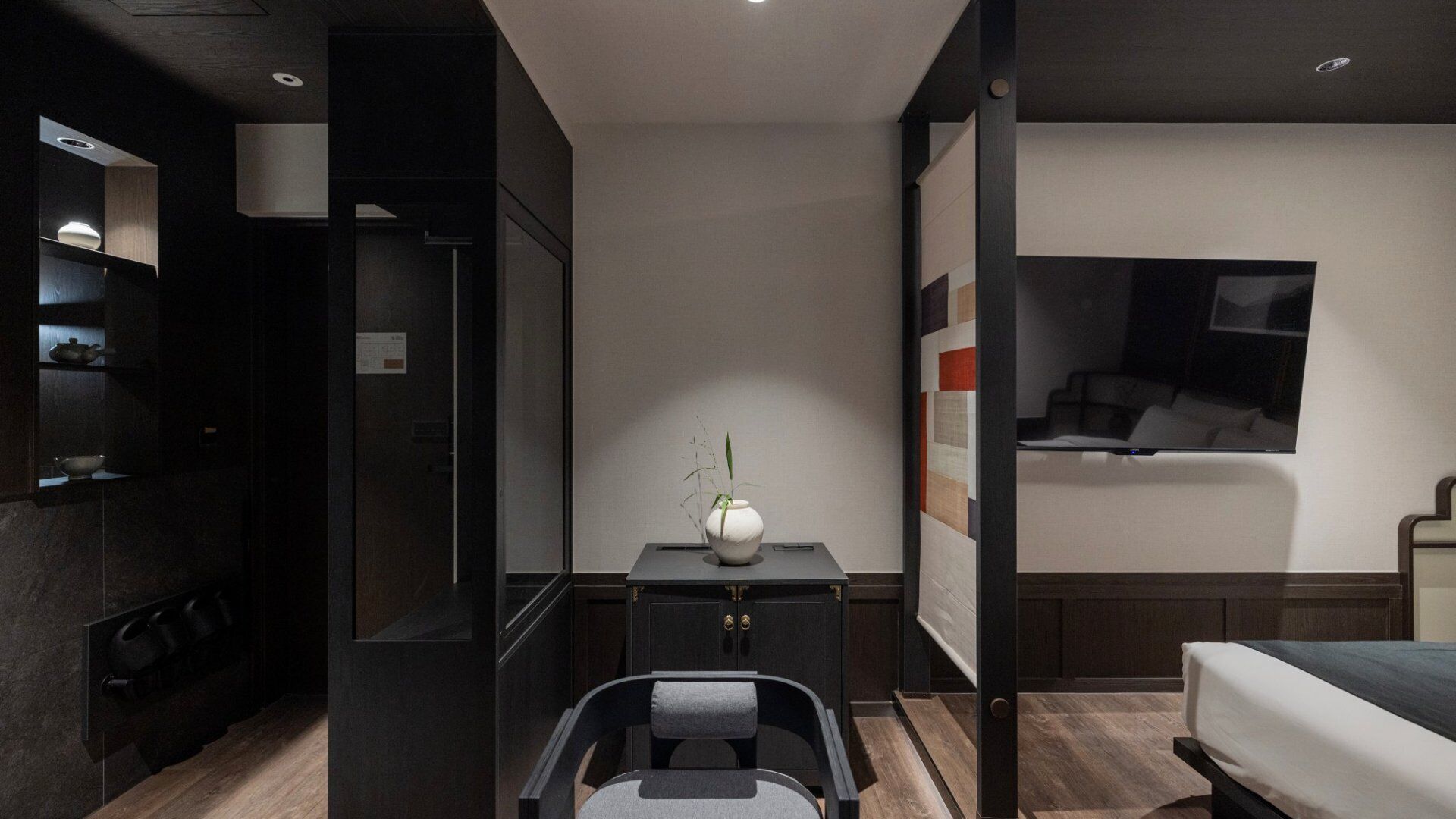 Quarto de hotel moderno com uma cama, uma escrivaninha com um vaso, uma cadeira, uma televisão e um guarda-roupa.