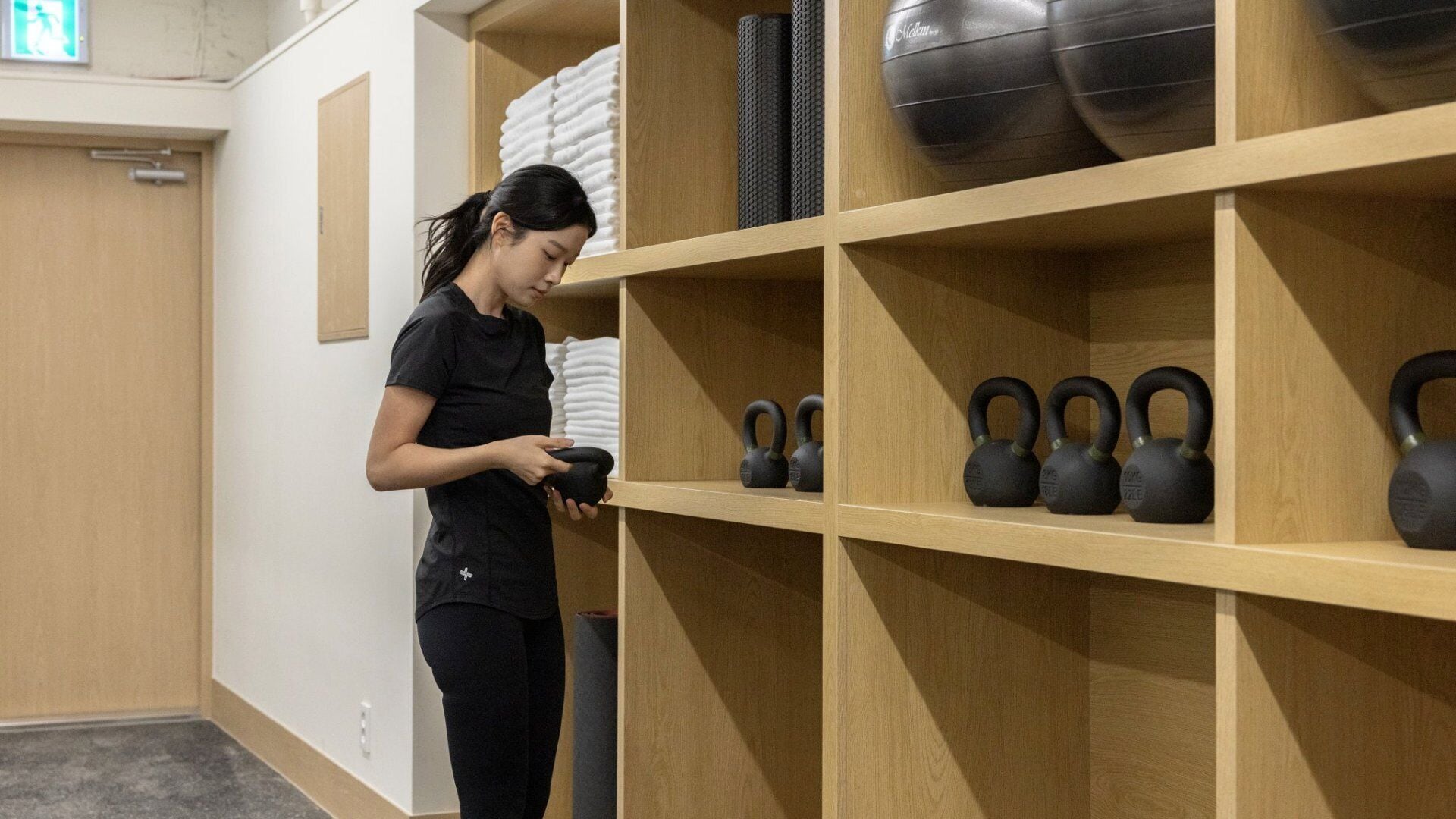 A person in a gym setting with wooden shelves containing kettlebells and folded towels.