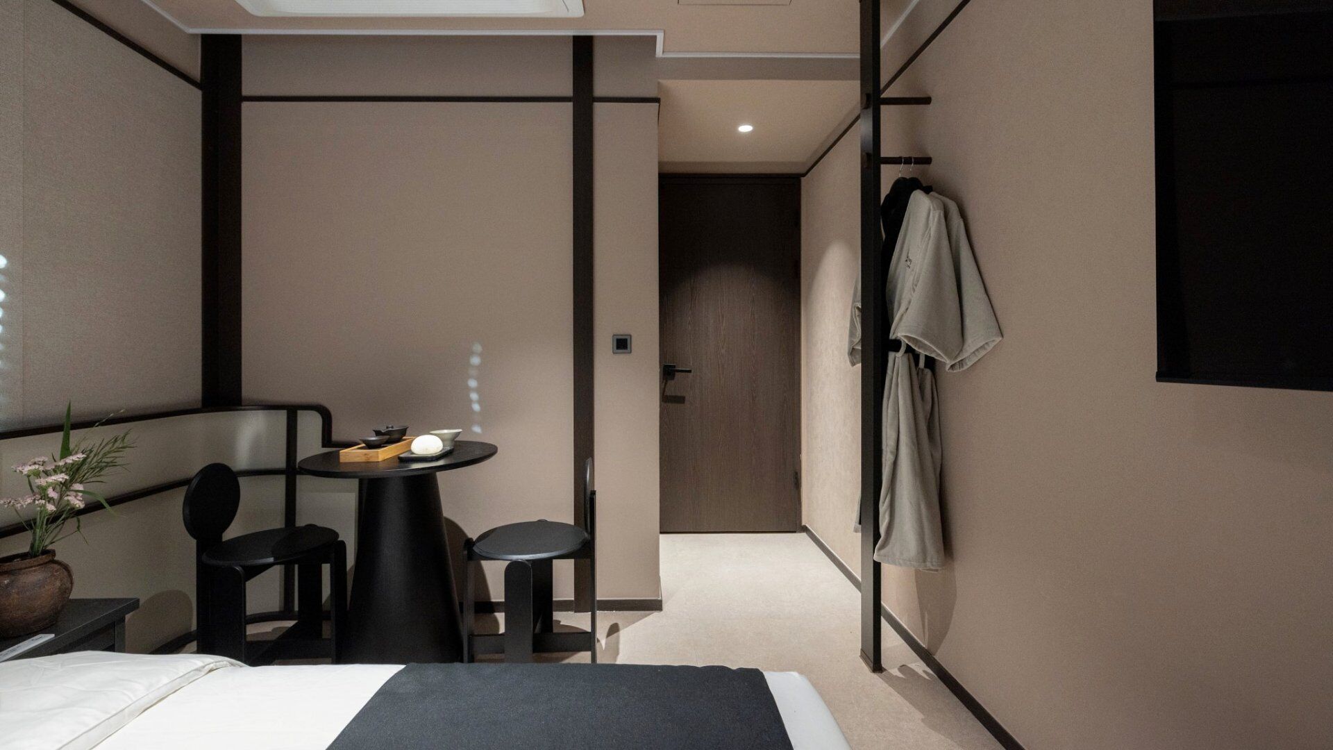 Quarto de hotel compacto com uma cama, uma mesinha com uma tigela e hashis, uma planta em vaso e um cabideiro com um casaco.