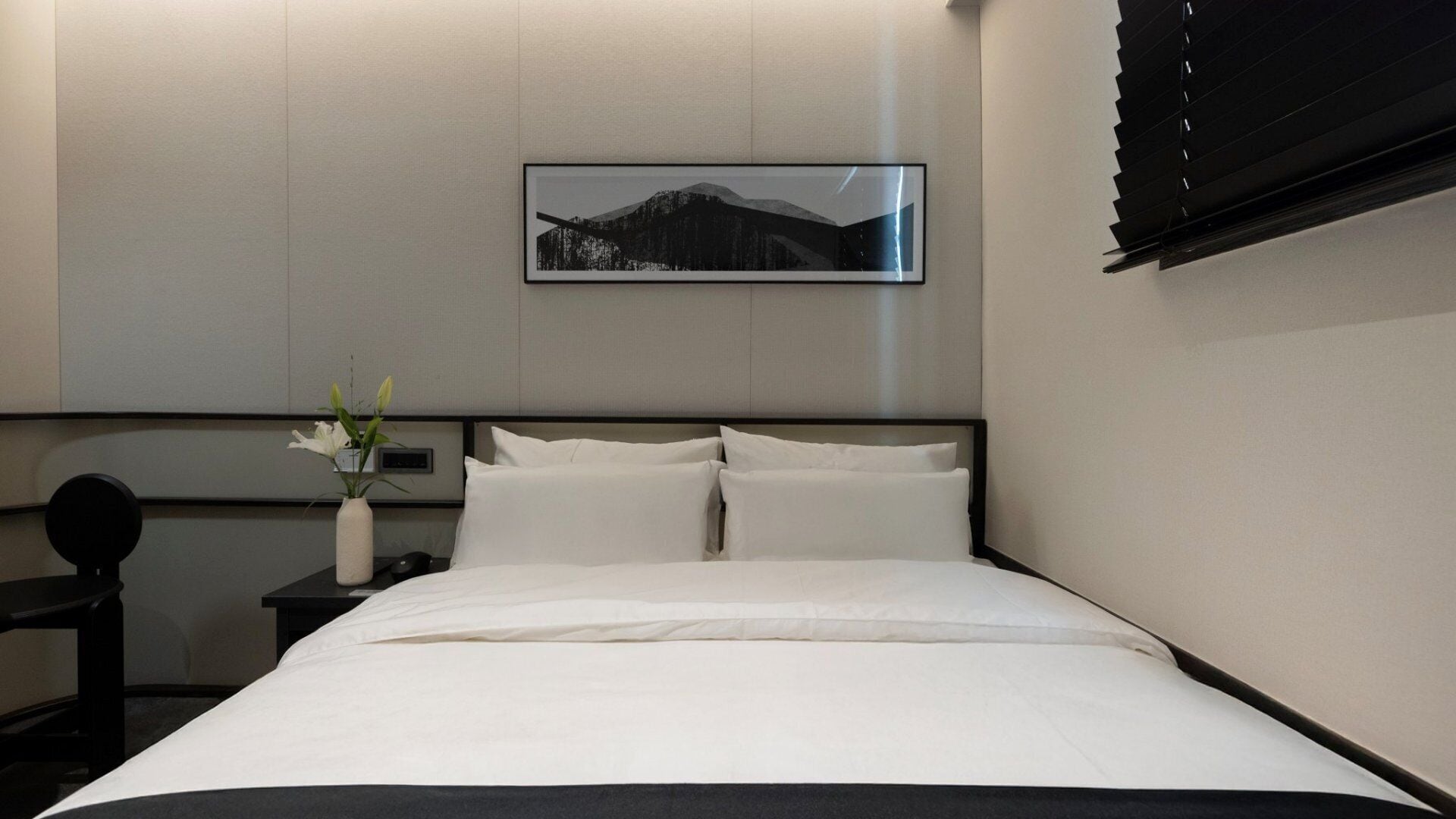Ein ordentlich bezogenes Bett mit weißen Laken, ein Nachttisch mit Blumenvase und ein gerahmtes Bild einer Berglandschaft an der Wand.