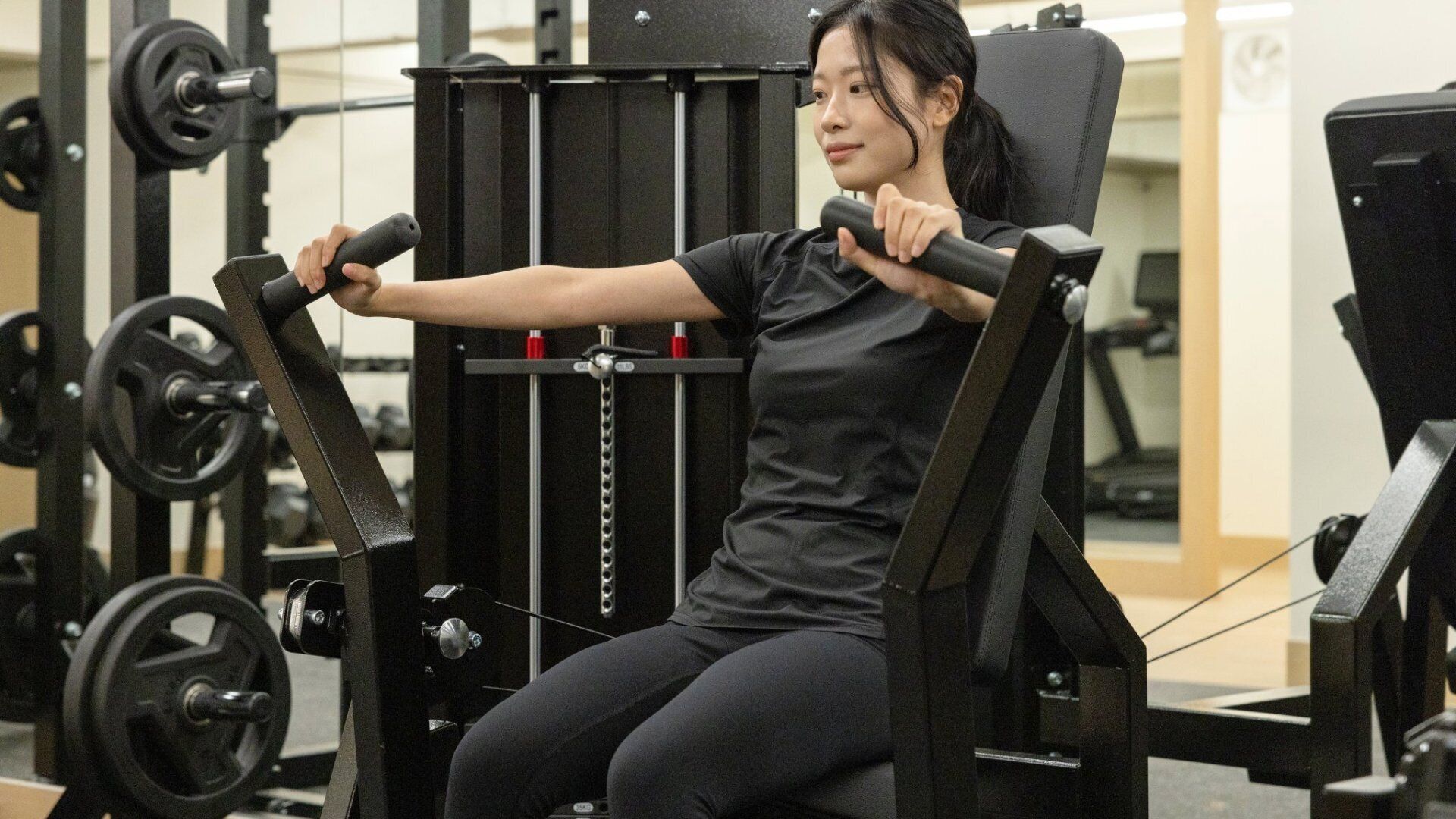 Uma pessoa usando uma máquina de leg press em uma academia, com diversos equipamentos de musculação ao fundo.