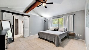 Ein Schlafzimmer mit einem Bett, Deckenventilator, einem Fenster mit Vorhängen und einer Tür zu einem anderen Raum.