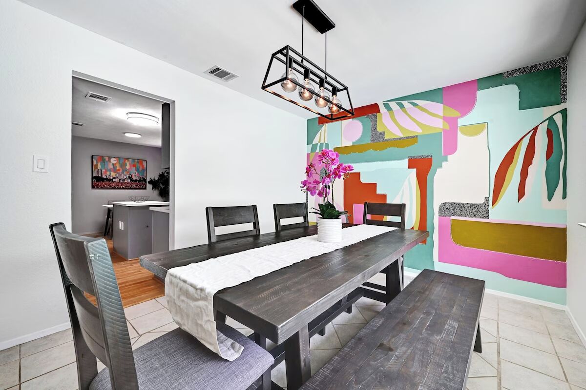 A dining area with a long wooden table, chairs, and a vase with flowers. There is a colorful abstract painting on the wall and a kitchen visible in the background.