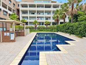 Una piscina con piso de baldosas, rodeada por un muro de ladrillo y un edificio con varios balcones.