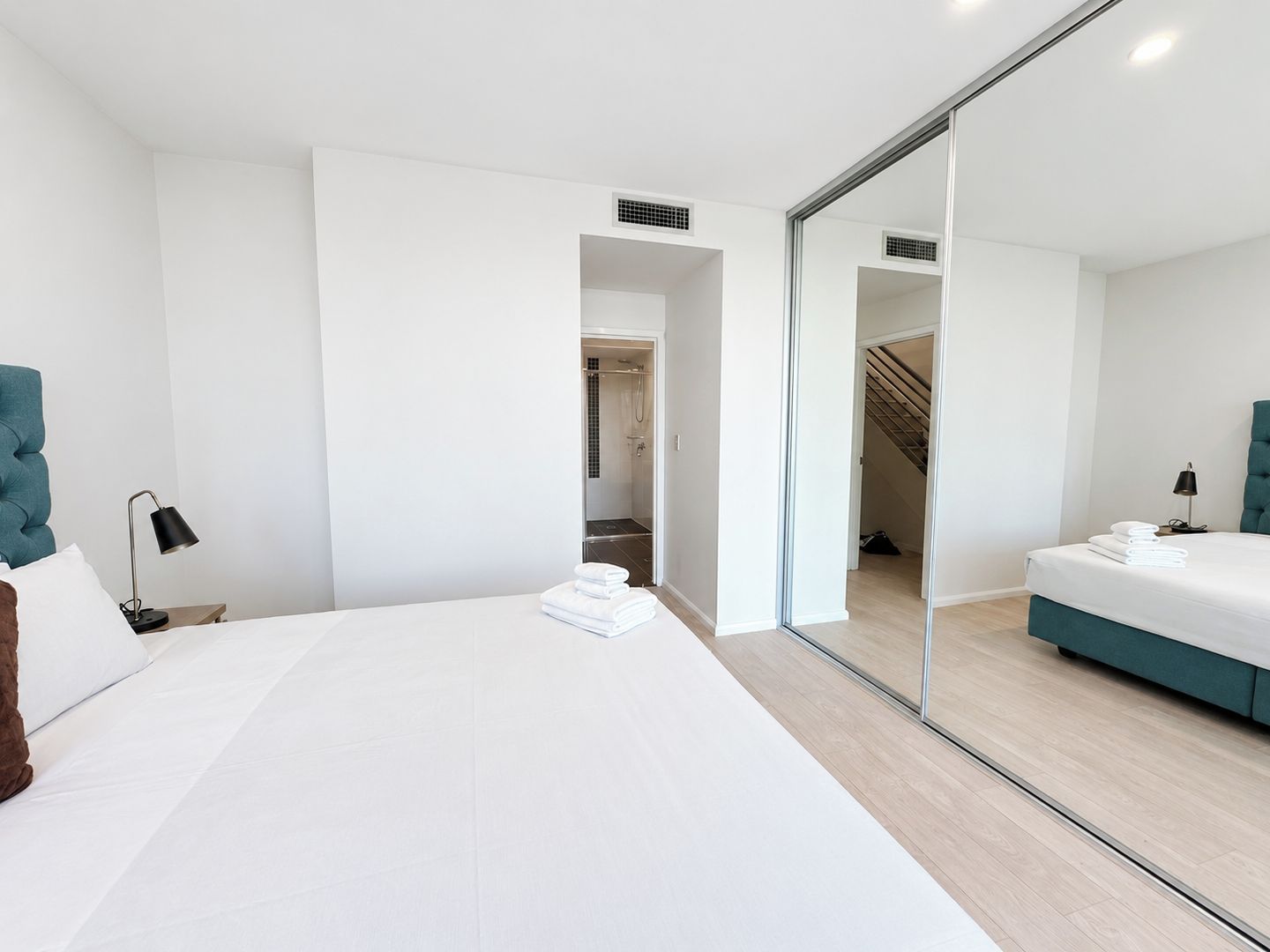 A modern hotel room with a large bed, a bedside table with a lamp, a mirror, and a view of a staircase.