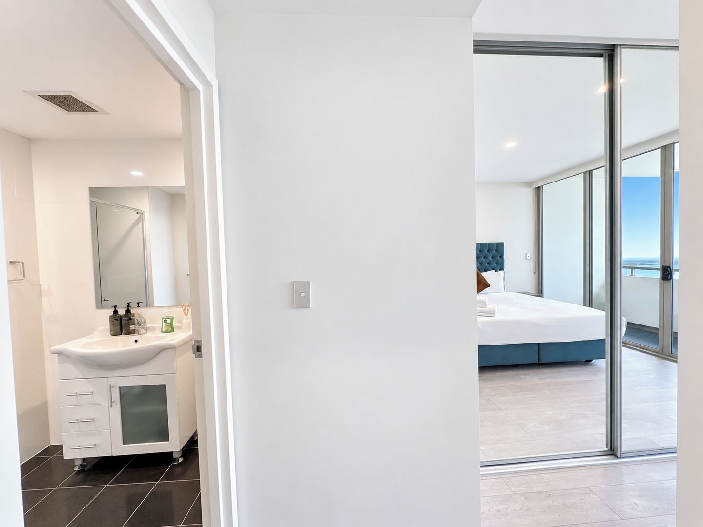 Un baño moderno con un lavabo y espejo blancos, y un dormitorio con una cama grande y vistas a través de un ventanal.