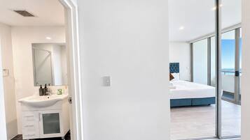 Un baño moderno con un lavabo y espejo blancos, y un dormitorio con una cama grande y vistas a través de un ventanal.