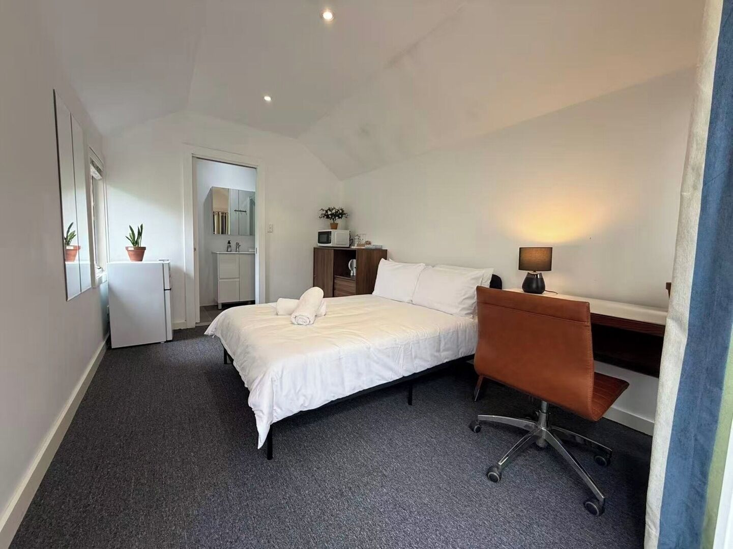 A hotel room with a bed, a desk, a chair, a small kitchenette, and a bathroom.