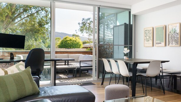 A modern living room with a sofa, a coffee table, and a TV. There is a dining area with a table and chairs. The room has large windows with a view of trees and hills. - Quiet Modern Apartment in Prime Location (Reid)