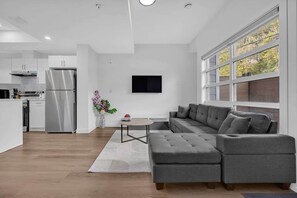 Une cuisine moderne avec un réfrigérateur en acier inoxydable, un coin repas avec un canapé gris et une petite table, et une grande fenêtre donnant sur les arbres.