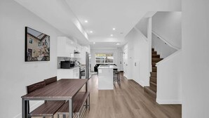 Una cocina moderna con armarios blancos, una zona de comedor con una mesa de madera y una escalera que sube a otro nivel.
