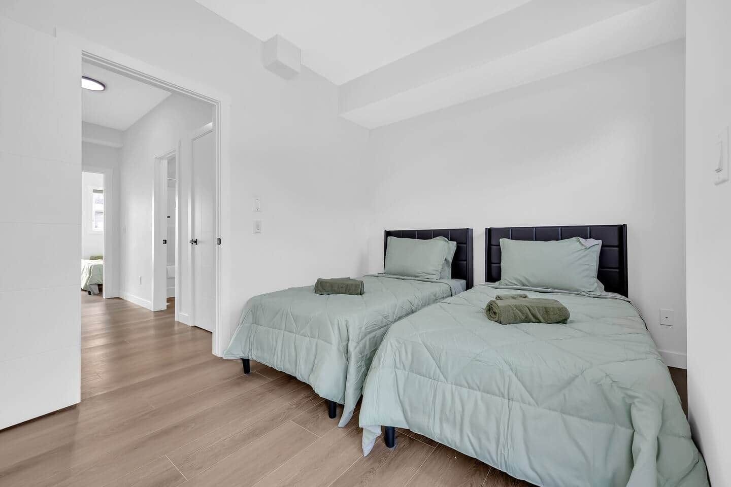 Un dormitorio con dos camas, piso de madera, paredes blancas y una puerta que da a otra habitación.