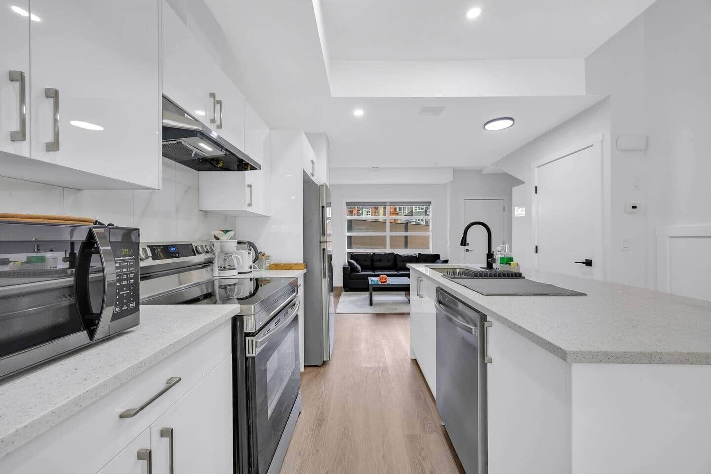 A modern kitchen with a microwave, oven, and sink, leading to a living area with a sofa and a window.